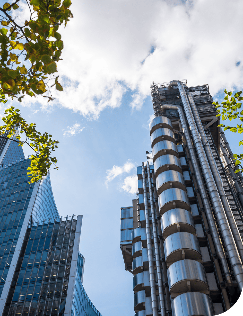 High-rise modern office buildings with glass facades and stainless steel industrial structures against a blue sky and trees, representing corporate construction and architectural design.